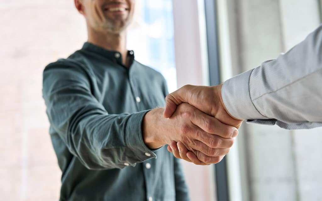 Two individuals standing by a window shaking hands, representing a successful merger and acquisition deal.