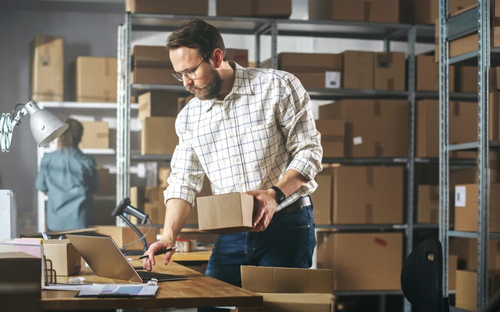 A warehouse worker inspects inventory on a laptop, holding a small box in one hand. Shelves filled with stacked boxes stretch into the background, illustrating the organized complexity of the storage system and highlighting the efficiency required for seamless warehouse operations.