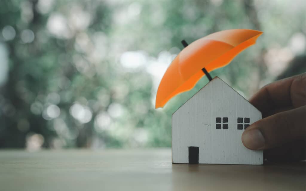 A small house figurine with a traditional design is sheltered under a bright orange umbrella, symbolizing protection and security. The blurred outdoor background features soft greens and browns, suggesting a natural setting. This composition conveys themes of home safety and insurance coverage.