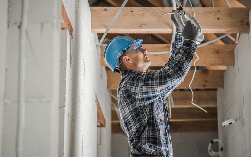 A construction worker, wearing a blue hard hat and plaid shirt, stands on a ladder while installing wiring on a ceiling. The worker is focused and uses both hands to securely position the wires. Dim lighting and exposed beams suggest an unfinished or industrial setting. The scene conveys precision and safety in a construction environment.