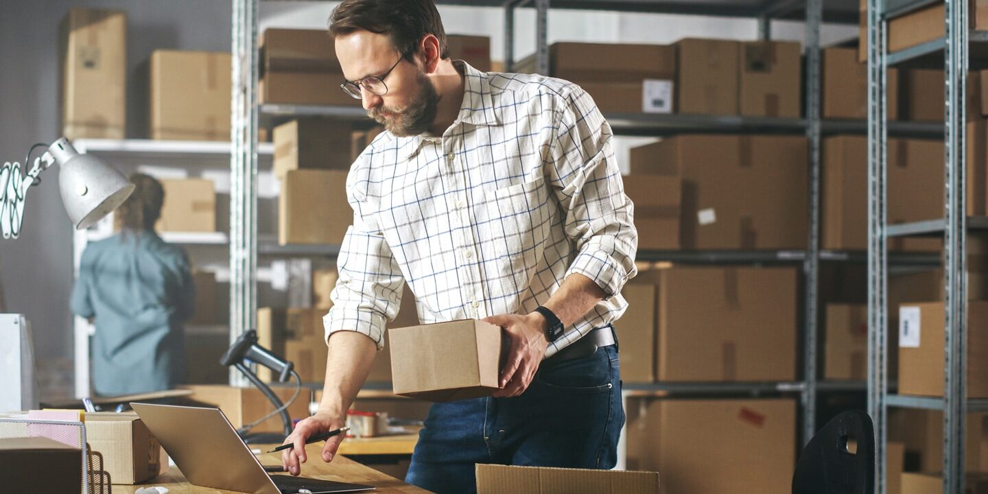 A warehouse worker inspects inventory on a laptop, holding a small box in one hand. Shelves filled with stacked boxes stretch into the background, illustrating the organized complexity of the storage system and highlighting the efficiency required for seamless warehouse operations.