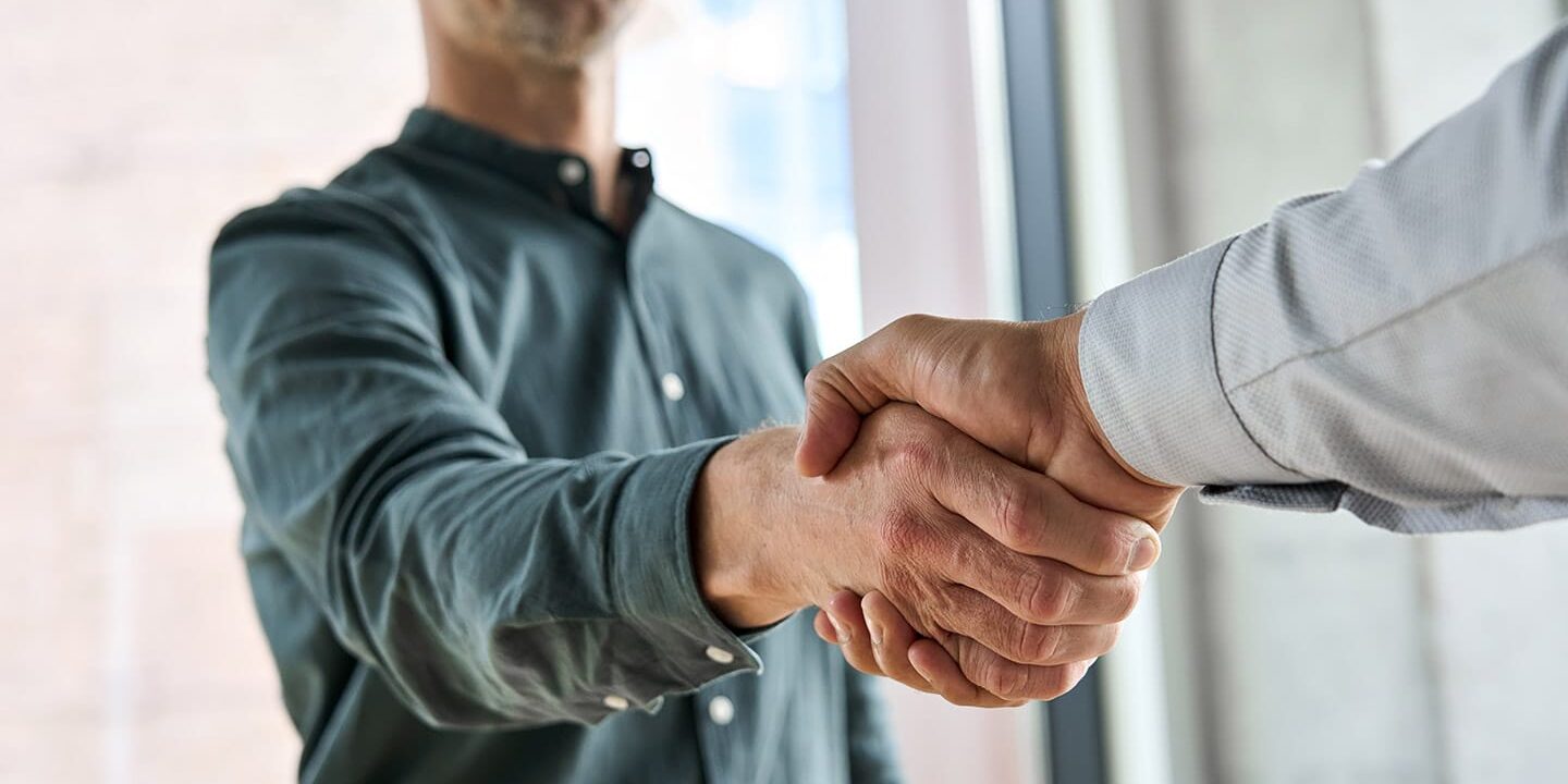 Two individuals standing by a window shaking hands, representing a successful merger and acquisition deal.